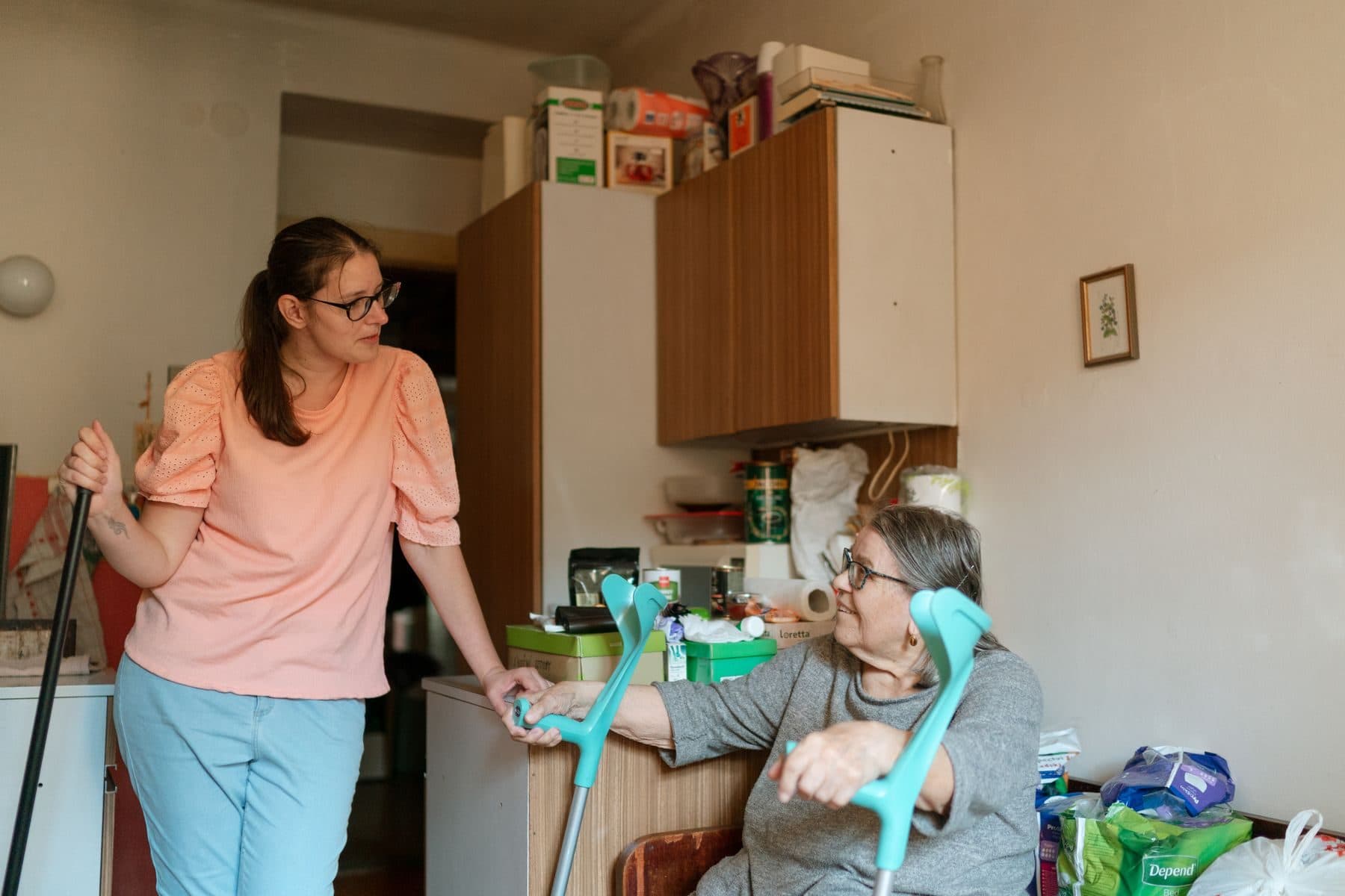 A carer supporting an older woman who uses crutches in her kitchen