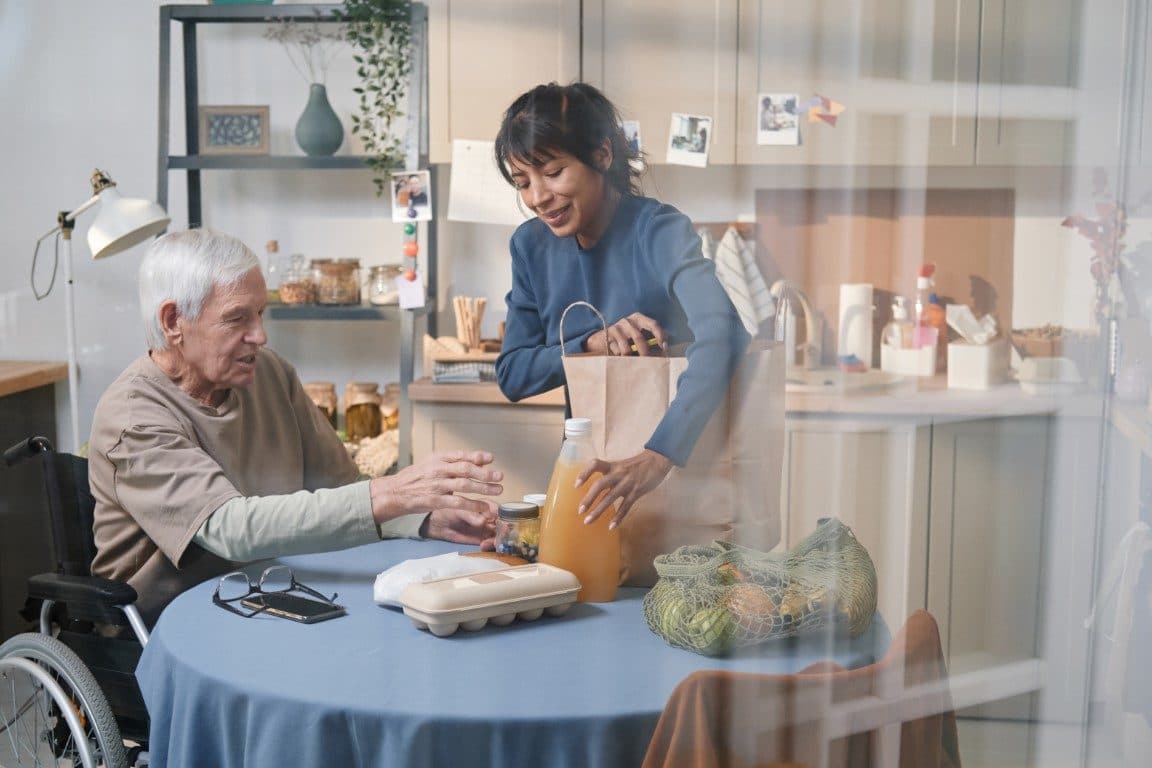 A carer unpacking shopping with an older man at his kitchen table